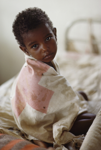 Foto van sterk vermagerd kind gewikkeld in een zak van het Rode Kruis, bij een voedselcentrum van het Rode Kruis in Ethiopië, gemaakt in 1986